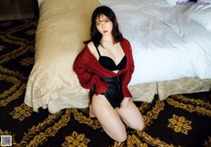 A woman sitting on a bed wearing a red shirt and black shorts.