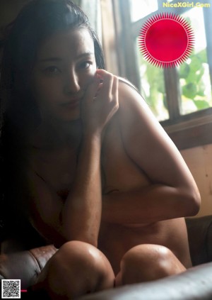 A naked woman sitting on a couch in front of a window.