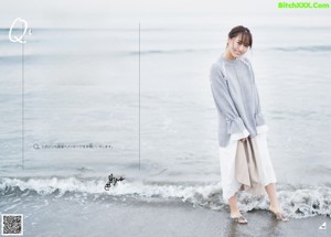 A woman standing on the beach wearing a gray sweater and white skirt.